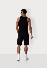 Schwarzes ärmelloses Tanktop, schwarze Jeansshorts mit ausgefranstem Saum, weiße Sneakers und knöchellange Socken; Tattoos sind auf dem Rücken sichtbar.