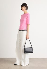 Vrouw in een roze top en brede witte broek, met een zwarte handtas met een schouderband, staande tegen een effen lichte achtergrond.