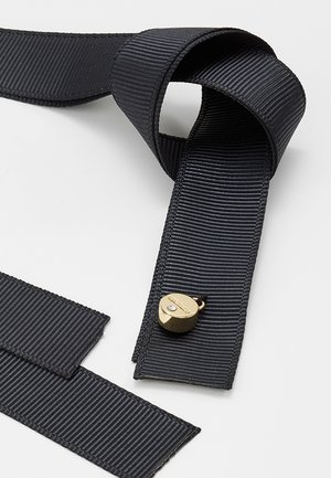 Sort ribbet stofrem med en lille lås i guldfarve, arrangeret i løse løkker på en lys grå overflade.