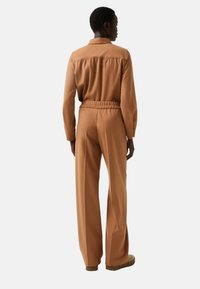 Bruine jumpsuit met lange mouwen, elastische tailleband en wijde benen, voorzien van een kraag en een details op de rug. Glad oppervlak.