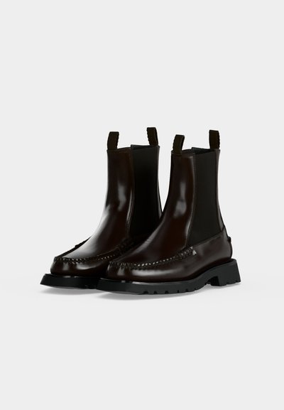 Paire de bottines en cuir marron foncé poli avec des semelles épaisses noires et des panneaux latéraux élastiques sur un fond clair uni.
