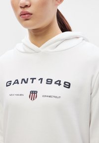 Hanorac alb cu grafică frontală cu "GANT1949" și un emblema de scut în roșu, alb și albastru. Material moale, croială lejeră și glugă cu șnur.