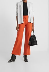 Cardigan gris léger, haut côtelé noir, pantalon large orange vif, bottes à talons en cuir noir, et sac seau en daim noir.