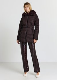 Braune Steppjacke mit Kunstpelzkapuze, tailliertem Schnitt und Reißverschlusstaschen, kombiniert mit glänzenden, dunkelbraunen Schlaghosen und weißen High Heels.