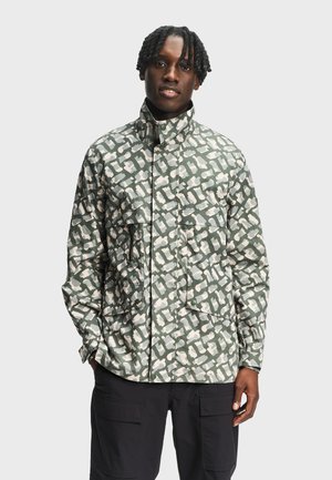 Homme portant une veste à motifs abstraits verts et beiges avec un col haut et un pantalon cargo noir, se tenant devant un fond clair uni.
