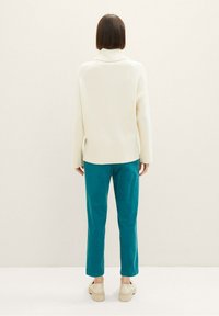 Personne vue de dos portant un pull à col roulé crème, un pantalon droit teal et des chaussures à plateforme beiges sur un fond uni.