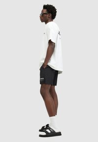 White oversized t-shirt, black shorts with logo, black sandals with adjustable straps, white crew socks. Model stands sideways against a gray background.