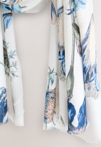 Foulard à motifs floraux en tissu léger ; présente des fleurs bleues et roses sur fond blanc, avec des bords doux et une texture subtile.