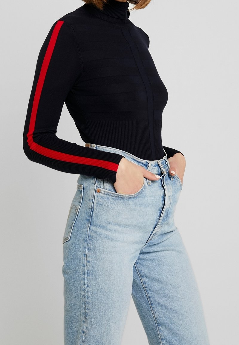 Personne portant un pull à col roulé noir avec des rayures rouges sur les manches et un jean taille haute bleu clair, mains dans les poches.