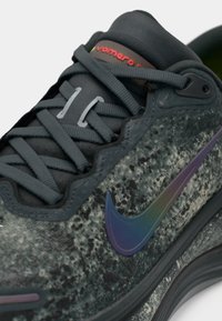 Nahaufnahme eines schwarz-grau gesprenkelten Sneakers mit einem dunklen, schimmernden Nike-Swoosh und gekreuzten schwarzen Schnürsenkeln auf weißem Hintergrund.