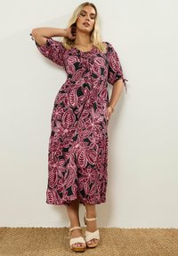 Frau im mid-length Kleid mit rosa Blumenmustern auf Schwarz, die mit einem Arm hinter ihrem Kopf posiert und auf einer strukturierten Matte vor einer hellen Wand steht.