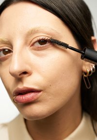 Brosse de mascara noir avec des poils fins tenue près de l'œil d'un modèle, arborant des sourcils pâles, un teint naturel et un subtil anneau labial doré.