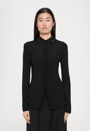 Schwarze gerippte Strickjacke mit Knopfleiste, langen Ärmeln und spitzem Kragen. Verfügt über einen kleinen Akzent am Kragen. Glatte Textur.