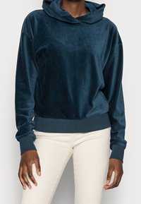 Marinblå velourhoodie med lös passform, over-sized huva och ribbade muddar och nederkant. Bärs över ljusfärgade byxor.