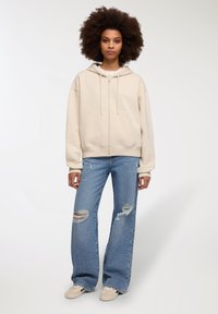 Beżowa bluza z zamkiem błyskawicznym, z kieszenią kangurką, zestawiona z jasnoniebieskimi, zniszczonymi spodniami typu wide-leg oraz jasnymi trampkami.