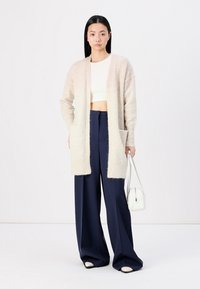 Cardigan aperto color beige chiaro realizzato in morbido tessuto a maglia, abbinato a un top bianco cropped e pantaloni a gamba larga blu scuro a vita alta.
