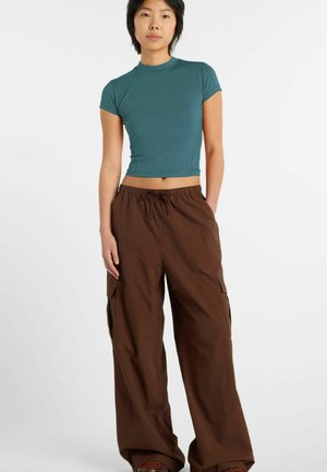 Donna che indossa un top corto aderente a maniche corte verde acqua e pantaloni cargo marroni larghi, in piedi contro uno sfondo bianco semplice.