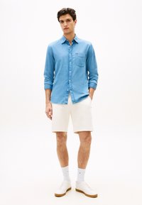 Ανοιχτό μπλε πουκάμισο από τζιν με κουμπιά και τσέπη στο στήθος, συνδυασμένο με λευκά σορτς και λευκά αθλητικά παπούτσια. Το πουκάμισο έχει μανίκια γυρισμένα και άνετη εφαρμογή.