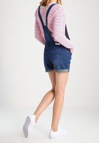 Denim overalls in dark blue with adjustable straps, paired with a red and white striped long-sleeve shirt. White sneakers complete the outfit.