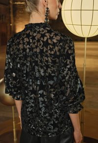 Blusa nera trasparente con motivo a stelle, tessuto testurizzato, scollatura arricciata e maniche voluminosi. La modella si trova vicino a una luce a forma di globo.