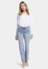 Camisa blanca de botones con detalle de pliegues, combinada con jeans rectos de color azul claro. El modelo lleva zapatos planos beige. Tejidos suaves y ligeros.