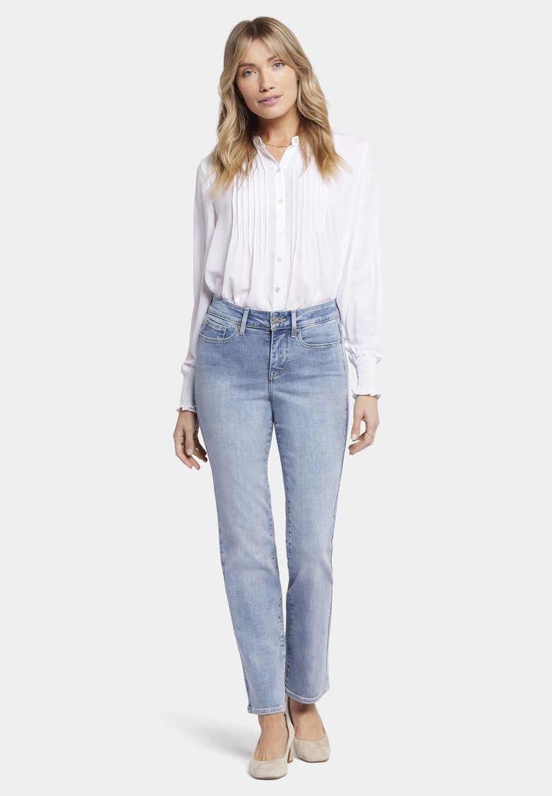 Camisa blanca de botones con detalle de pliegues, combinada con jeans rectos de color azul claro. El modelo lleva zapatos planos beige. Tejidos suaves y ligeros.