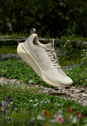 Světlé béžové běžecké boty značky Nike zavěšené nad štěrkovou cestou s zelenou trávou a divokými květinami v pozadí.