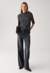 Femme se tenant debout, portant un haut noir à motifs, un jean gris foncé taille haute à jambes larges, des chaussures noires, tenant un parapluie noir plié.
