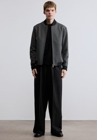 Γκρι μπουφάν τύπου bomber με μαύρες λεπτομέρειες, φερμουάρ μπροστά και ριμπ μανσέτες, συνδυασμένο με φαρδιά μαύρα παντελόνια· λείο ύφασμα και μινιμαλιστικός σχεδιασμός.