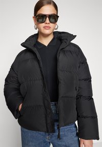 Donna che indossa un giubbotto imbottito nero, un polo scuro, jeans blu, grandi orecchini a cerchio argentati e occhiali da sole rettangolari oversize, con le mani nelle tasche.