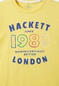 Żółta bawełniana koszulka z napisem "HACKETT" w wyraźnych niebieskich literach oraz "1983" w zielonym, czerwonym i białym, z napisem "QUINTESSENTIALLY BRITISH LONDON" poniżej.