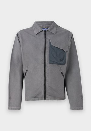 Pelēka jaka ar apkakli, priekšējo rāvējslēdzēju un divām sānu kabatām. Iezīmējas ar lielu, struktūru apveltītu kabatu kreisajā krūtīs. Kokvilnas materiāls.