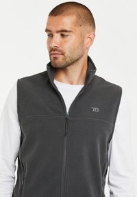 Grau Fleece-Weste mit hohem Kragen, Frontreißverschluss und Logo-Akzent. Getragen über einem weißen Langarmshirt, mit seitlichen Taschen.