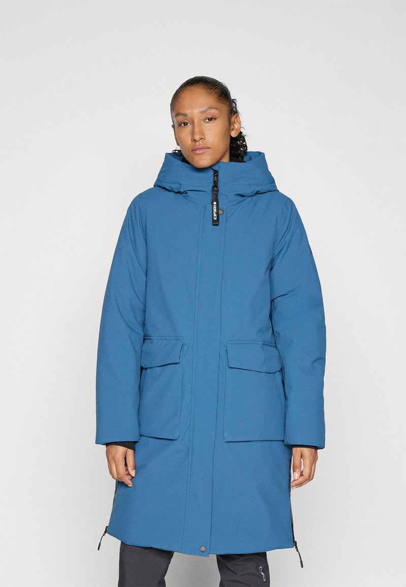 Frau in einem langen blauen Wintermantel mit Kapuze und vorderen Klapptaschen, die vor einem schlichten hellen Hintergrund steht.