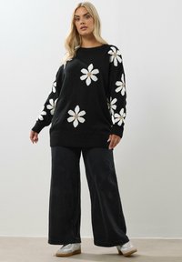 Schwarzer Pullover mit weißen Gänseblümchenmustern und beigen Zentren, kombiniert mit weit geschnittenen schwarzen Hosen und weißen Sneakers. Kuschelige Textur und lässiges Design.