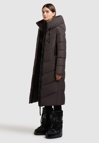 Lang mørkebrun pufferjakke med skrå quiltet mønster, oversized hætte, frontlynlås og sidelommer. Sammen med sorte vinterstøvler.