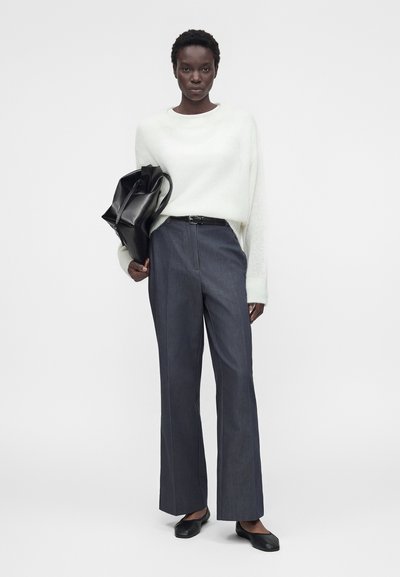 Γυναίκα με λευκό πουλόβερ, σκούρο παντελόνι με φαρδιά μπατζάκια, μαύρα φλατ παπούτσια, κρατώντας μεγάλη μαύρη δερμάτινη τσάντα μπροστά σε απλό λευκό φόντο.