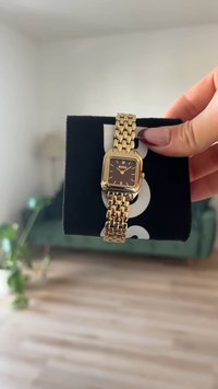 Gold-toned wristwatch with a square face, dark brown dial, gold links, and Roman numerals. Displayed on a black box.