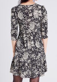 Robe noire à fleurs avec des manches trois-quarts, présentant un motif floral blanc, une jupe évasée et un col rond. Texture de tissu douce.
