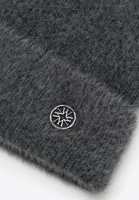 Grijze gebreide muts van zachte wol, voorzien van een gestructureerde afwerking en een ronde zilveren embleem met een geometrisch ontwerp.