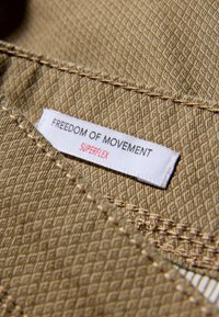 Beiger Stoff mit strukturiertem Muster. Das Etikett trägt die Aufschrift "FREEDOM OF MOVEMENT" und "SUPERFLEX" in schwarzer und roter Schrift. Sichtbare Nähte.