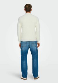 Krämfärgad ribbad tröja i kombination med blå cargobyxor med flera fickor, avslappnad passform och vita sneakers. Enfärgad bakgrund.