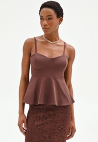 Mujer con top marrón sin mangas con peplum y falda a juego de encaje floral, complementada con un collar grueso de cadena dorada.