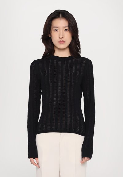 Pull en tricot noir à manches longues avec des rayures verticales, une texture côtelée et un col rond, assorti à un pantalon de couleur claire.