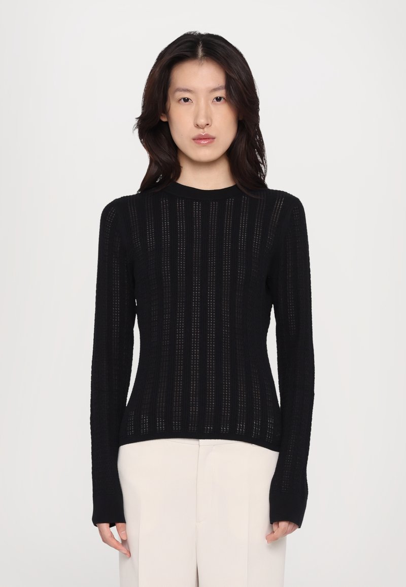 Sort langærmet strikket sweater med lodrette striber, ribbet tekstur og rund hals, parret med lyse bukser.