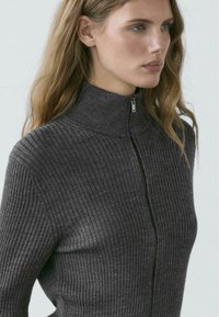 Grauer Rippenpullover mit hohem Kragen und durchgehendem Reißverschluss, mit strukturiertem Strickmuster und figurbetonter Silhouette.