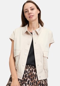 Beżowa, krótka kurtka z krótkimi rękawami, z zapięciem na guziki i dwiema przednimi kieszeniami. Stworzona na bazie czarnej bluzki i wzorzystej spódnicy.
