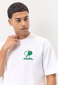 Maglietta bianca in cotone con logo verde "PADEL" e grafica di una racchetta da tennis e una pallina sull'area del petto sinistro. Maniche corte, design a girocollo.