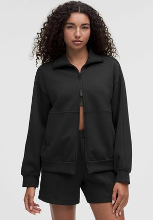Vrouw met krullend haar draagt een zwart jack met rits en bijpassende zwarte korte broek, staand tegen een eenvoudige lichte achtergrond.
