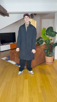 Lang svart frakk over en beige topp, svarte bukser og sølvfargede sko. Plante og brun sofa i bakgrunnen. Parkettgulv med mønstret teppe.